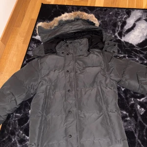 Grå canada goose whydnham black label - Säljer en grå dunjacka från Canada Goose med svart logotyp på ärmen. Jackan har huva med fuskpäls, stora fickor och stängs med både dragkedja och knappar. Perfekt för kalla dagar och har en klassisk, stilren look.