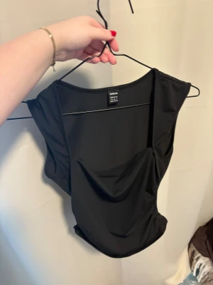 Svart topp  - Säljer en svart, stilren topp från SHEIN i storlek S. Toppen har en cool draperad detalj framtill och har en tight passform. Perfekt att styla med jeans eller kjol för en trendig look. Materialet är mjukt och stretchigt syntet.