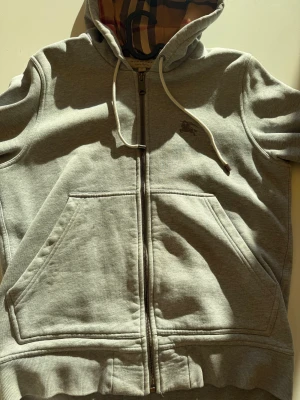 Grå Burberry hoodie med dragkedja Äkta  - Snygg grå hoodie från Burberry med klassiskt rutigt foder i huvan och längs insidan. Tröjan har dragkedja, snörning vid huvan och två stora fickor framtill. Materialet är mjuk bomull och polyester, perfekt för chill dagar. Ikonisk Burberry-logga broderad på bröstet.