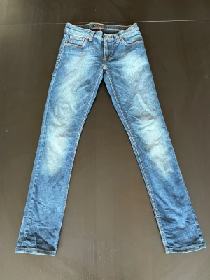 Nudie Tight Long John W26 L32  - Säljer ett par blå skinny jeans från Nudie Jeans med klassisk orange söm och broderad bakficka. Jeansen har smal passform, normal midja och är tillverkade i mjukt denimtyg. Perfekta för dig som gillar en slimmad look och snygga detaljer.