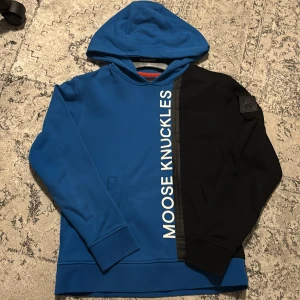 Blå och svart hoodie Moose Knuckles - Snygg hoodie från Moose Knuckles i blått och svart med stor vit logga längs framsidan. Tröjan har huva, lång ärm och en cool patch på ena ärmen. Materialet är mjukt och känns som bomull och polyester. Perfekt för dig som vill sticka ut med streetstyle.