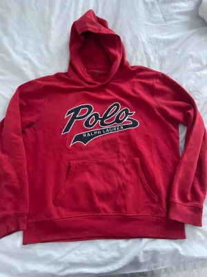 Röd hoodie från Polo Ralph Lauren - Säljer en röd hoodie från Polo Ralph Lauren med stor logotyp på bröstet. Tröjan har huva, magficka och långa ärmar. Tillverkad i mjuk bomull som är skön att ha på sig. Perfekt för dig som gillar streetwear och klassisk stil.