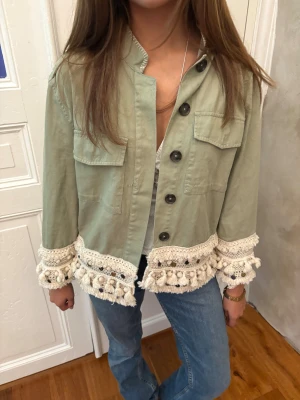 Grön jeansjacka med virkade detaljer - Snygg ljusgrön overshirt med stora svarta knappar och två bröstfickor. Jackan har unika virkade detaljer med tofsar i beige längs nederkant och ärmslut som ger boho-vibe. Perfekt att styla med jeans för en chill look.