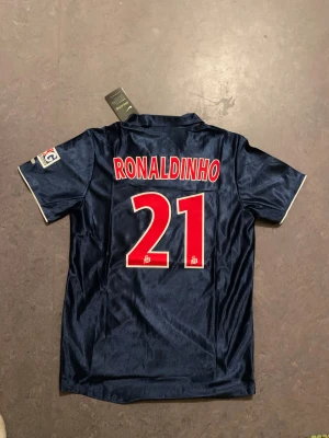 PSG Ronaldinho 21 Nike fotbollströja - Säljer en klassisk Paris Saint-Germain fotbollströja med Ronaldinho och nummer 21 på ryggen. Tröjan är marinblå med en röd vertikal rand, vita detaljer och PSG-logga på bröstet. Tillverkad i glansigt polyester, har krage och korta ärmar. Perfekt för dig som älskar retro och fotbollshistoria.