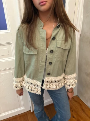 Grön jeansjacka med virkade detaljer - Snygg ljusgrön overshirt med stora svarta knappar och två bröstfickor. Jackan har unika virkade detaljer med tofsar i beige längs nederkant och ärmslut som ger boho-vibe. Perfekt att styla med jeans för en chill look.