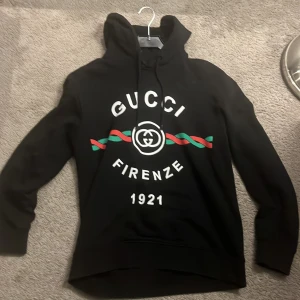 Svart Gucci hoodie med tryck - Svart hoodie från Gucci med stor logotyp och texten 'Firenze 1921' på bröstet. Snyggt tryck i vitt, rött och grönt över magen. Tröjan har huva med snörning och långa ärmar. Perfekt för dig som gillar exklusiva streetwear-vibbar.