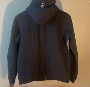 Svar  fleecejacka Sail Racing - Mörkblå fleecejacka från Sail Racing med huva och grov dragkedja i metall. Jackan har lång ärm, ficka med dragkedja på bröstet och broderad logga på både huva och bröst. Perfekt för kyliga dagar och snygg till jeans. 