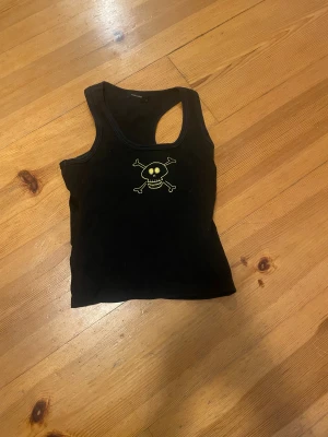 Svart linnetopp med döskalle Vero Moda - Cool svart linnetopp från Vero Moda i storlek S. Toppen har ett tryck med en gul döskalle och korslagda ben på bröstet. Modellen är ärmlös med rundad halsringning och tillverkad i mjuk bomull. Perfekt för dig som gillar edgy stil!