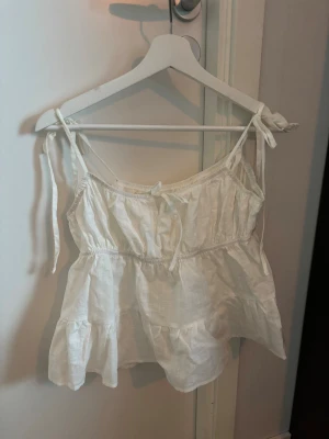 Vit linnetopp med volanger och knyt - Supersöt vit linnetopp med volanger och knytband på axlarna. Toppen har en luftig passform, volangkant nedtill och en liten rosett framtill. Materialet är lätt och känns som linne, perfekt för varma dagar. Axelbanden kan justeras med knyt för extra komfort.