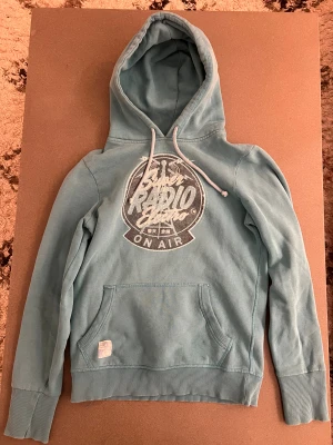 Ljusblå Superdry hoodie med tryck - Snygg ljusblå hoodie från Superdry med stort vitt tryck på bröstet och coola detaljer. Tröjan har huva med snörning, känguruficka och ribbade muddar. Perfekt för dig som vill ha en chill look.🤩😸