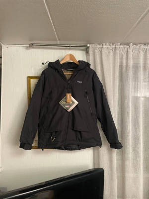 Svart Patagonia vinterjacka med huva! - Svart vinterjacka från Patagonia med huva och dragkedja framtill. Jackan är tillverkad i Gore-Tex-material, har två stora fickor med dragkedja och justerbara muddar. Längd 67cm Armlängd 57 cm +/-1cm Goretex material så den ska tåla regn!