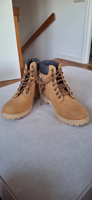 Bruna boots från FILA med snörning - Snygga bruna boots från FILA i mockaimitation med grov sula och snörning. Skorna har svart vadderad kant upptill för extra komfort och en robust design som passar perfekt till höst- och vinteroutfits. Klassisk rund tå och diskret FILA-logga på sidan.