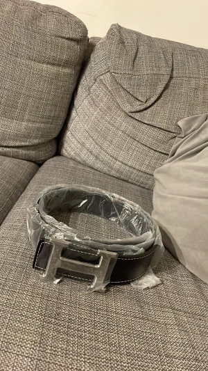 Svart skärp med H-spänne - Snyggt svart bälte med ett stort H-format metallspänne och vita kontrastsömmar längs kanterna. Bältet har en stilren och modern look, perfekt för att lyfta din outfit. Materialet är skinn och spännet är i silverfärgad metall.