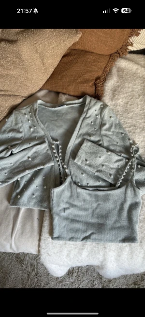 Ljusgrå croppatt set med pärlor från Zara - Superfin ljusgrå croppad topp från Zara med matchande cardigan. Båda delarna har pärldetaljer och är ribbade. Toppen är ärmlös med smala axelband och cardigannen har långa ärmar och knyt framtill. Perfekt för dig som gillar stilrena och söta detaljer. Färg turkos