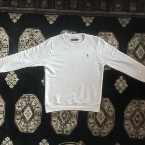 Vit sweatshirt från Polo Ralph Lauren - Vit sweatshirt från Polo Ralph Lauren med klassisk rund hals och broderad logga på bröstet. Tröjan är tillverkad i 100% bomull och har långa ärmar samt ribbade muddar vid hals, ärmslut och nederkant. Perfekt för chill dagar och enkel att matcha.