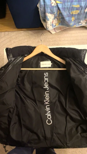 Svart pufferjacka från Calvin Klein Jeans - Säljer en svart, glansig pufferjacka från Calvin Klein Jeans med dragkedja framtill och hög krage. Jackan har en clean look med logga på bröstet och insidan, samt vadderade paneler som ger extra volym. Perfekt för dig som gillar streetstyle och vill hålla dig varm. Den är i storlek 16 men passar mig som vanligtvis har S hade också kunnat passa XS, mer bilder finns så skriv ifall du vill ha de!💕 pris kan diskuteras!