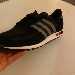 Adidas LA Trainer svarta sneakers - Adidas LA Trainer sneakers i svart med gråa ränder och detaljer. Snygg retrostil med mesh och mocka på ovansidan, samt klassisk platt sula med färgade pluggar i hälen. Helt ny och aldrig använd. Kommer direkt ifrån boxen. Toppenskick! Storlek 42! Passar både på vintern och sommarn!