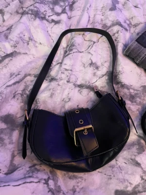 Svart axelväska med bälte-detalj - Snygg svart axelväska i skinnimitation med cool bälte-detalj och guldfärgade metalldelar. Väskan har rundad form och justerbar axelrem, perfekt för att ge din outfit en trendig touch. Enkel att matcha och rymmer det viktigaste du behöver ha med dig.