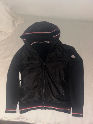 Svart Moncler bomberjacka med huva - Svart bomberjacka från Moncler med huva och dragkedja. Jackan har ribbade muddar med röd och vit rand, samt Moncler-logga på ärmen. Framsidan är quiltad och har två fickor med dragkedja. Materialmix av polyester och bomull.