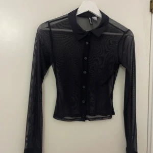 Svart meshskjorta från H&M Divided XS - Svart transparent meshskjorta från H&M Divided i storlek XS. Skjortan har klassisk krage, långa ärmar och knappar framtill. Snygg slim passform och lätt material som ger en cool vibe. Perfekt att styla lager på lager eller med ett linne/bh under.