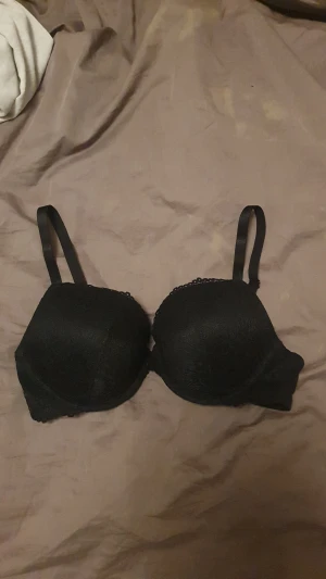 Svart BH I Spets - Säljer en svart bh med vadderade kupor och justerbara axelband. Bhn har ett snyggt spetsmönster över hela ytan och en feminin kant upptill. En liten kudde i upptäckte jag när jag kom hem därav säljer jag.