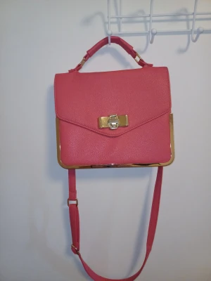 Rosa axelväska med gulddetaljer - OBS KOLLA NOGA PÅ 2 OCH 3 BILDEN OCH PÅ SWSTA SÅ SER MAN INTE NEN DET FINNS NÅN ENSTAKA LITE FLÄK MEN MAN SER KNAPT.Snygg rosa axelväska med guldfärgade metalldetaljer och ett elegant lås framtill. Väskan har både handtag och band så man kan ha den över axen, samt ett rymligt fack med dragkedja på insidan. Perfekt för dig som vill ha en stilren och din väska ❤️ 