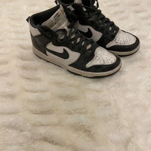 Nike panda dunks High svart/vit sneakers - Nike Dunk High sneakers i svart och vitt med klassisk swoosh på sidorna. Skorna har snörning, hög siluett och perforerad tåbox. Ovandelen är i skinn och textil, med svart foder och vit plös. Perfekt för dig som gillar streetstyle och retro vibes.