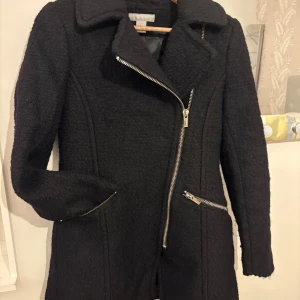 Svart ullkappa med dragkedjor - Stilren svart kappa i ullmix med markerad krage och snedställd dragkedja framtill. Två sneda fickor med dragkedjor ger en edgy vibe. Fodrad insida i glansigt tyg. Perfekt för dig som gillar en clean och modern look.