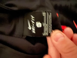 Svart Nike hoodie Dri-Fit XL - Svart hoodie från Nike i storlek XL med Dri-Fit material som andas. Snyggt tryck i rött framtill med ZS-logga och Nike swoosh. Långärmad modell med huva, perfekt för träning eller chill. Materialet är lätt och funktionellt.