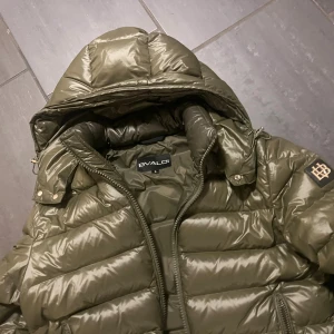 Olivgrön pufferjacka från Bvaldi - Säljer en olivgrön pufferjacka från Bvaldi med huva och dragkedja. Jackan har quiltad design, två sidofickor med dragkedja och en snygg logotyp på ärmen. Perfekt för kalla dagar och riktigt skön att bära.