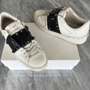 Valentino Rockstud Untitled sneakers - Snygga vita Valentino Rockstud Untitled sneakers med svart band och ikoniska nitar. Skorna har rund tå, snörning och coola detaljer längs sidorna och sulan. Tillverkade i skinn för en lyxig känsla och edgy look. Perfekta för dig som vill sticka ut med designerstil.