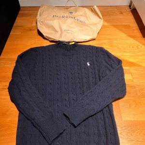  Polo Ralph Lauren stickad tröja - Kabelstickad mörkblå tröja från Polo Ralph Lauren med klassisk rund hals och broderad vit logga på bröstet. Tröjan har lång ärm och ribbade muddar för en snygg passform. Perfekt för dig som gillar stilren och tidlös design. Pris kan diskuteras!!