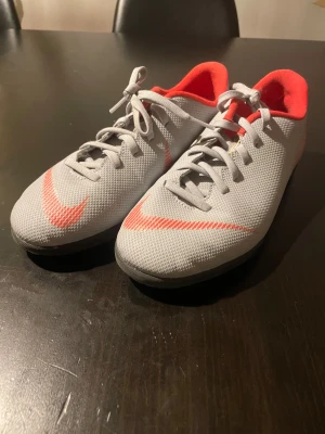 Grå Nike inomhussko med orange detaljer - Säljer ett par grå Nike inomhussko med snygga orange swoosh-detaljer och matchande orange insida. Skorna har snörning och är tillverkade i syntetmaterial med en platt sula som passar perfekt för spel inomhus. Riktigt stilrena och sportiga skor för dig som gillar inomhussport