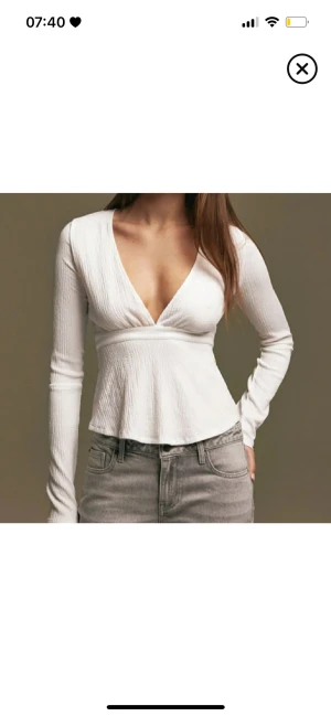 Vit långärmad omlottopp - Snygg vit långärmad topp med v-ringning och omlottkänsla. Toppen har en lätt strukturerad yta och markerad midja som ger en feminin siluett. Perfekt att styla med jeans eller kjol för en trendig look.