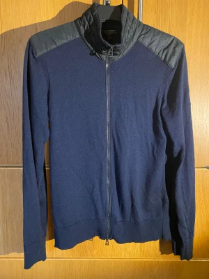 Mörkblå kofta från Belstaff med quiltad axel - Snygg mörkblå kofta från Belstaff i storlek M. Koftan har quiltade detaljer i svart på axlar och krage, samt hel dragkedja framtill. Stickad i mjukt material och med långärmad passform. Perfekt för dig som gillar stilrena och coola plagg.