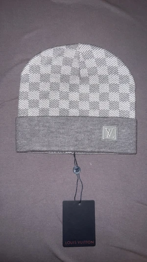 Grå rutig mössa Louis Vuitton - Snygg grå mössa från Louis Vuitton med klassiskt rutigt mönster och LV-logga på uppviket. Mössan är stickad och har en modern, clean look som passar perfekt till streetwear. Materialet känns mjukt och lyxigt.