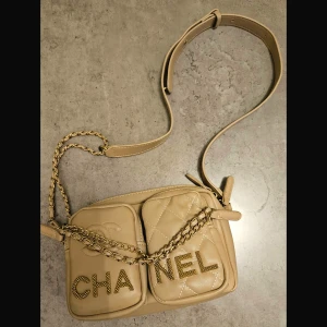 Beige axelväska - Säljer en beige axelväska med två framfickor, varav en är quiltad och båda har logotypen i guldfärgade nitar. Väskan har en justerbar axelrem i läder och guldfärgad kedjedetalj. Kontakta för fler bilder eller snabb affär :)