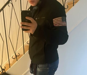 Svart vindjacka från Ralph Lauren - Svart vindjacka från Denim & Supply Ralph Lauren med amerikansk flaggpatch på ärmen, två bröstfickor med dragkedja och klassisk ståkrage. Jackan är tillverkad i polyester. Perfekt för dig som gillar street och sportig stil.              Passar storlek xs då den är verkligen liten i storleken 