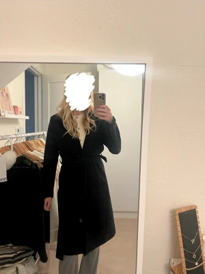 Svart lång kappa med bälte - Snygg svart kappa med klassisk krage och knappar hela vägen framtill. Modellen är lång och har ett matchande bälte i midjan. 