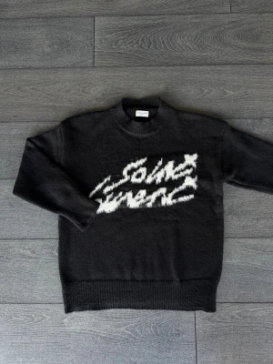 Svart Saint Laurent stickad tröja - Svart stickad tröja från Saint Laurent med vit, fluffig logotyp framtill. Tröjan har rund hals, långa ärmar och ribbade muddar. Den är gjord i mjukt material som känns lyxigt och har en oversized vibe. Perfekt för dig som gillar statement-plagg och designer.
