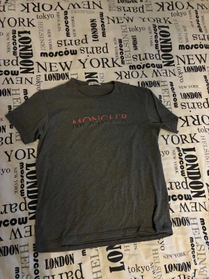 Grå Moncler t-shirt med tryck - Snygg grå t-shirt från Moncler i storlek S. T-shirten har ett stilrent tryck med Moncler-loggan i rött och svart på bröstet. Klassisk rund hals och korta ärmar, perfekt för en chill och enkel look. Materialet känns mjukt och bekvämt mot huden.