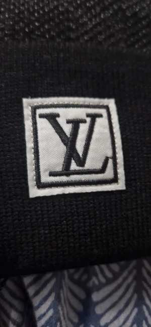 Svart rutig mössa med L v-logga - Svart mössa från L v med diskret rutmönster och uppvikt kant. Framtill sitter en vit patch med svart L v-logga. Mössan är stickad och har en exklusiv känsla, perfekt för dig som vill ha en clean och lyxig look.