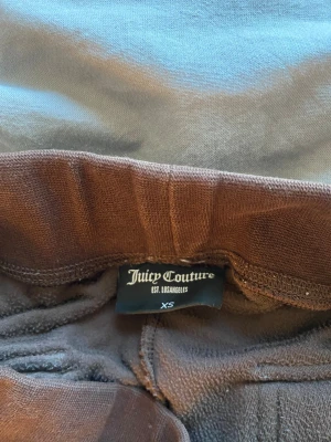 Bruna mjukisbyxor Juicy Couture XS - Bruna mjukisbyxor från Juicy Couture i storlek XS. Byxorna har bred resår med snörning i midjan, stora fickor fram och bak med knappar, samt broderad logga på ena fickan. Materialet är mjukt och skönt, perfekt för chill dagar.