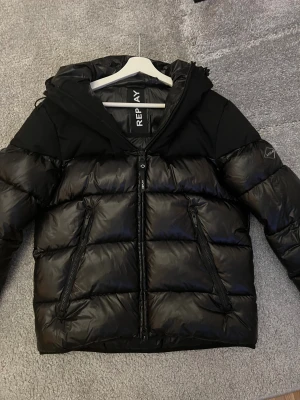 Svart pufferjacka från Replay - Snygg svart pufferjacka från Replay med huva och stora logotypdetaljer. Jackan har quiltad design, två dragkedjeförsedda fickor och en rejäl dragkedja framtill. Materialet är glansigt och känns riktigt modernt, perfekt för kalla dagar.