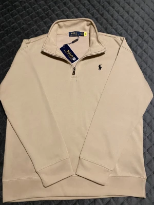 Beige half zip tröja Polo Ralph Lauren - Snygg beige långärmad tröja från Polo Ralph Lauren med klassisk svart logobrodyr på bröstet. Tröjan har en stilren ståkrage med half zip-dragkedja. Perfekt för dig som gillar clean och tidlös stil.