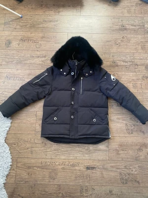 Svart dunjacka Moose Knuckles - Svart dunjacka från Moose Knuckles med pälskant på huvan, flera dragkedjor och knappar framtill. Jackan har ett snyggt metallmärke på ärmen och broderad etikett på insidan. Perfekt för kalla dagar med sin tjocka och puffiga design. Priset kan diskuteras.