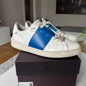 Valentino Open sneakers vit/blå - Säljer ett par Valentino Open sneakers i vitt skinn med en bred blå läderpanel på sidan och klassiska nitar runt hälen. Skorna har rund tå, platt sula och snörning. Perfekt för dig som gillar lyxiga och stilrena sneakers med en unik touch. Dustbag och snören medföljer. Tar även byten! Hör av er vid frågor ✅