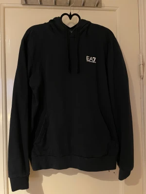 Svart hoodie EA7 Emporio Armani - Svart hoodie från EA7 Emporio Armani med vit logga på bröstet. Hoddien har huva med snörning, stor magficka och långa ärmar.  I bra skick och säljs i befintlig skick. 