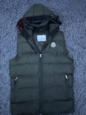 Mörkgrön dunväst från Moncler - Snygg mörkgrön dunväst från Moncler med huva och broderad logga på bröstet. Västen har två fickor med dragkedja och quiltad design. Perfekt för att hålla värmen och samtidigt se cool ut. Moncler-tryck på insidan av huvan ger extra stilpoäng.