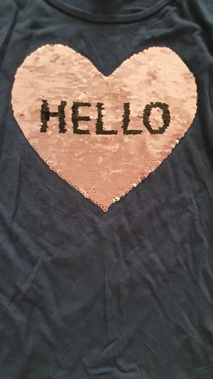 Tröja - Snygg mörkblå långärmad tröja från Lindex med stort hjärta i rosa paljetter på bröstet. Paljetterna kan vändas och visar antingen texten 'HELLO' eller ett par söta ögonfransar. Mjuk bomullskvalitet och rund hals. Perfekt för dig som gillar lite extra glitter.